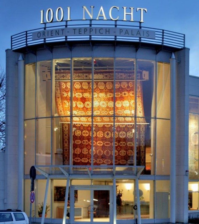 Der Rundbau des Orient-Teppich-Palais 1001 Nacht mit beleuchtetem Schriftzug und großem Orientteppich im Schaufenster